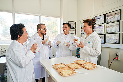 Four people in white lab coats taste noodles with chopsticks