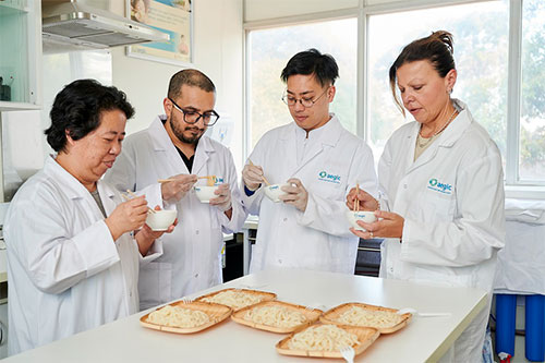 Four people in white lab coats taste noodles with chopsticks