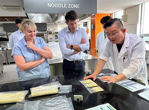 Three people stand around a counter with packets of noodles in bags in front of them