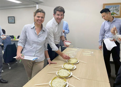 Two people stand by a table with plates of noodles, smiling at the camera