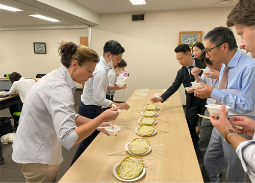 A group of people stand on both sides of a table with a number of plates of noodles in the middle