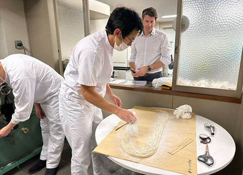 A man makes noodles on a table watched by another man through an open window